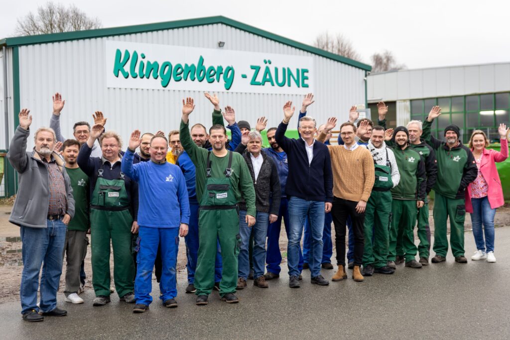 Das freundliche Team von Klingenberg Zaeune winkt vor der Werkshalle und signalisiert Kompetenz für professionelle Zaunmontage.